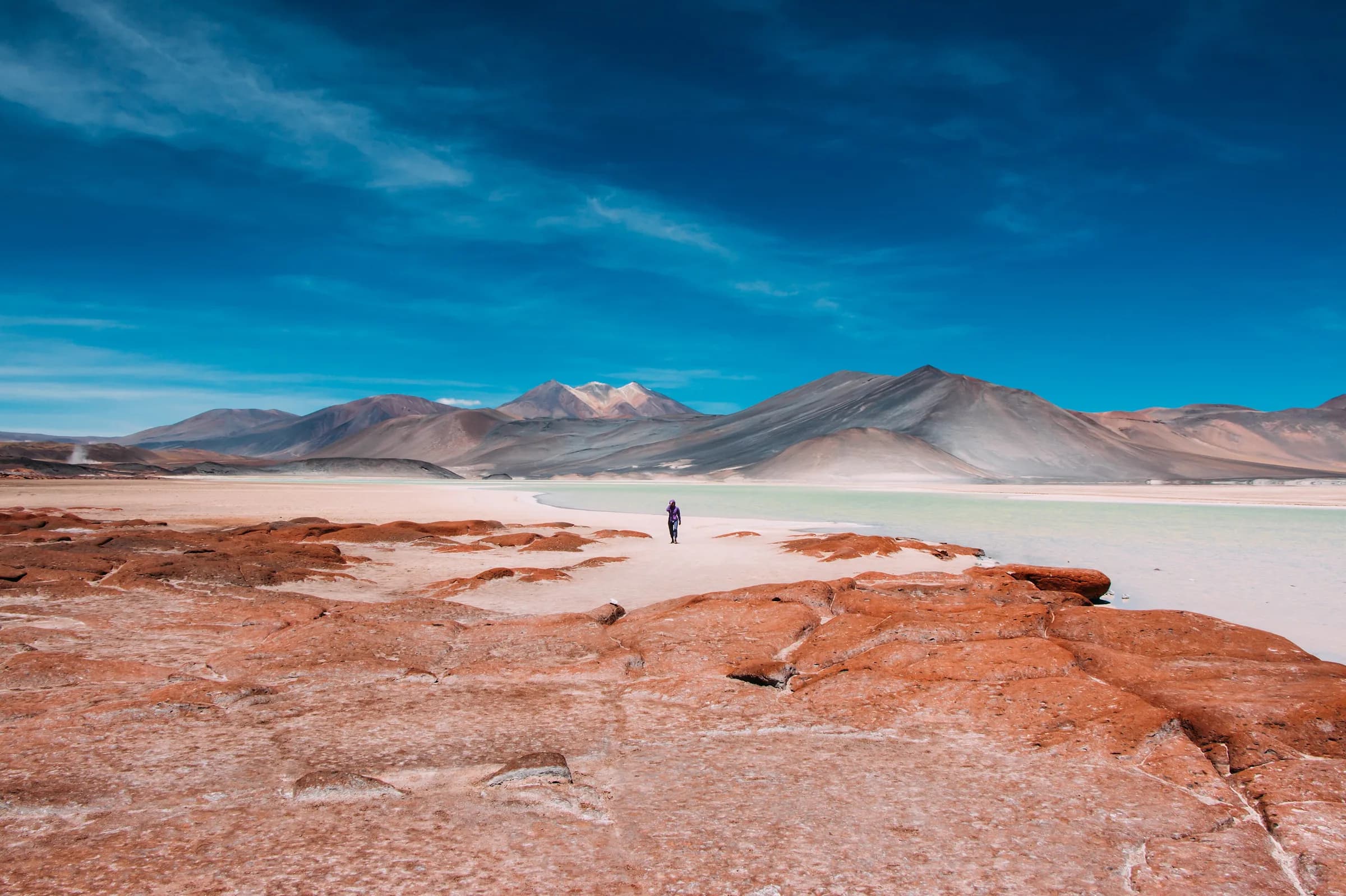 A traveler crossing red volcanic rock beside pale water and high Andes at Piedras Rojas in Chile's Atacama Desert