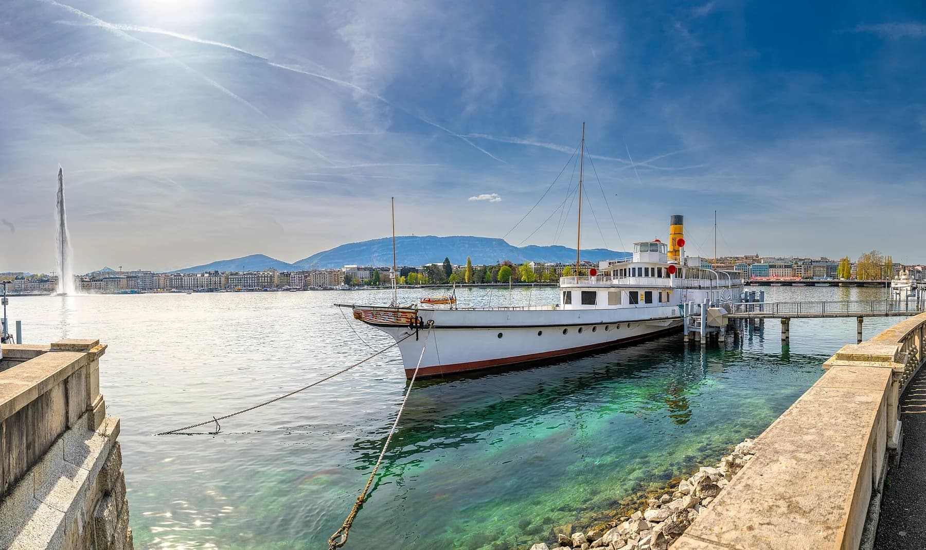 Lake Geneva harbor with clear water, a passenger boat, and mountains beyond the city